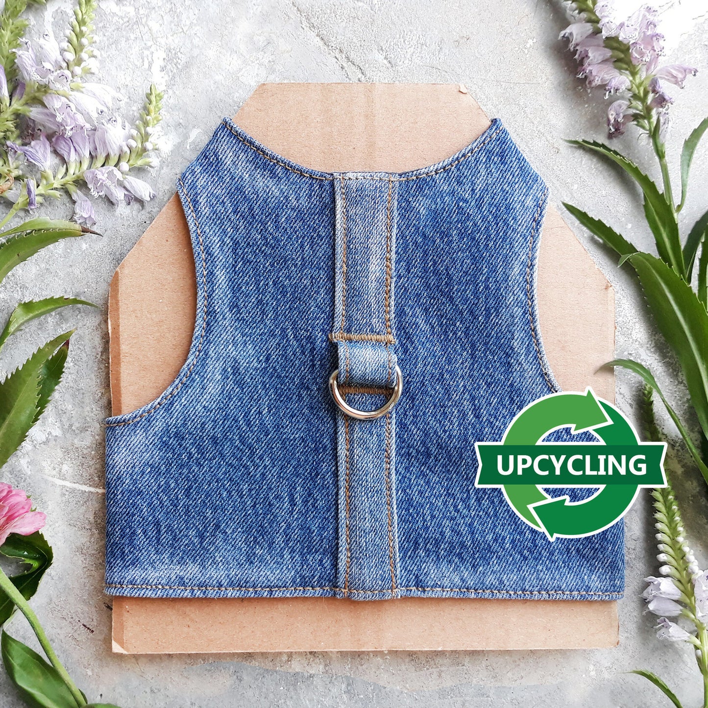 A cat harness made from blue denim fabric, featuring a central strap and a silver-colored D-ring.