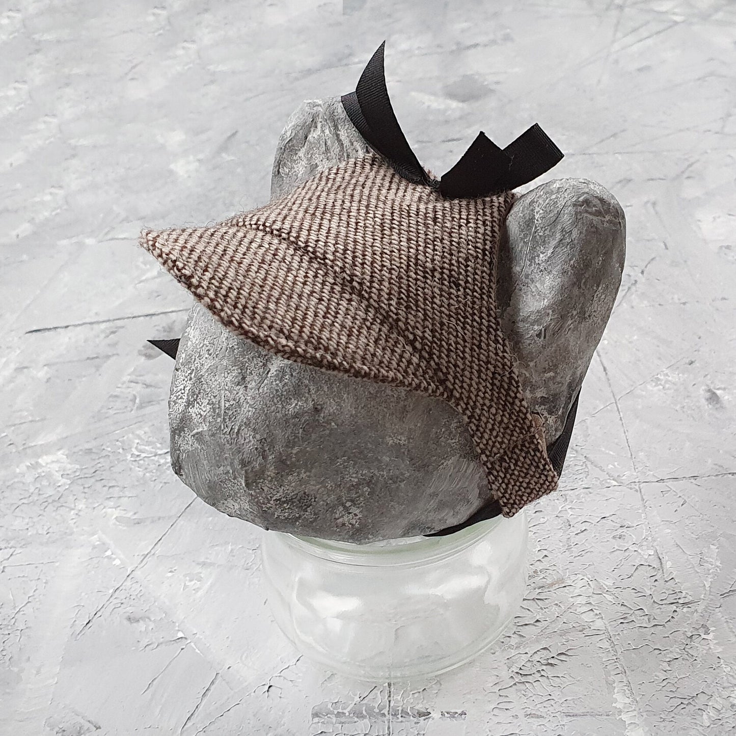Tweed-style hat for a cat with a small brim and a black ribbon tied in a bow on top, shown on a grey textured surface.