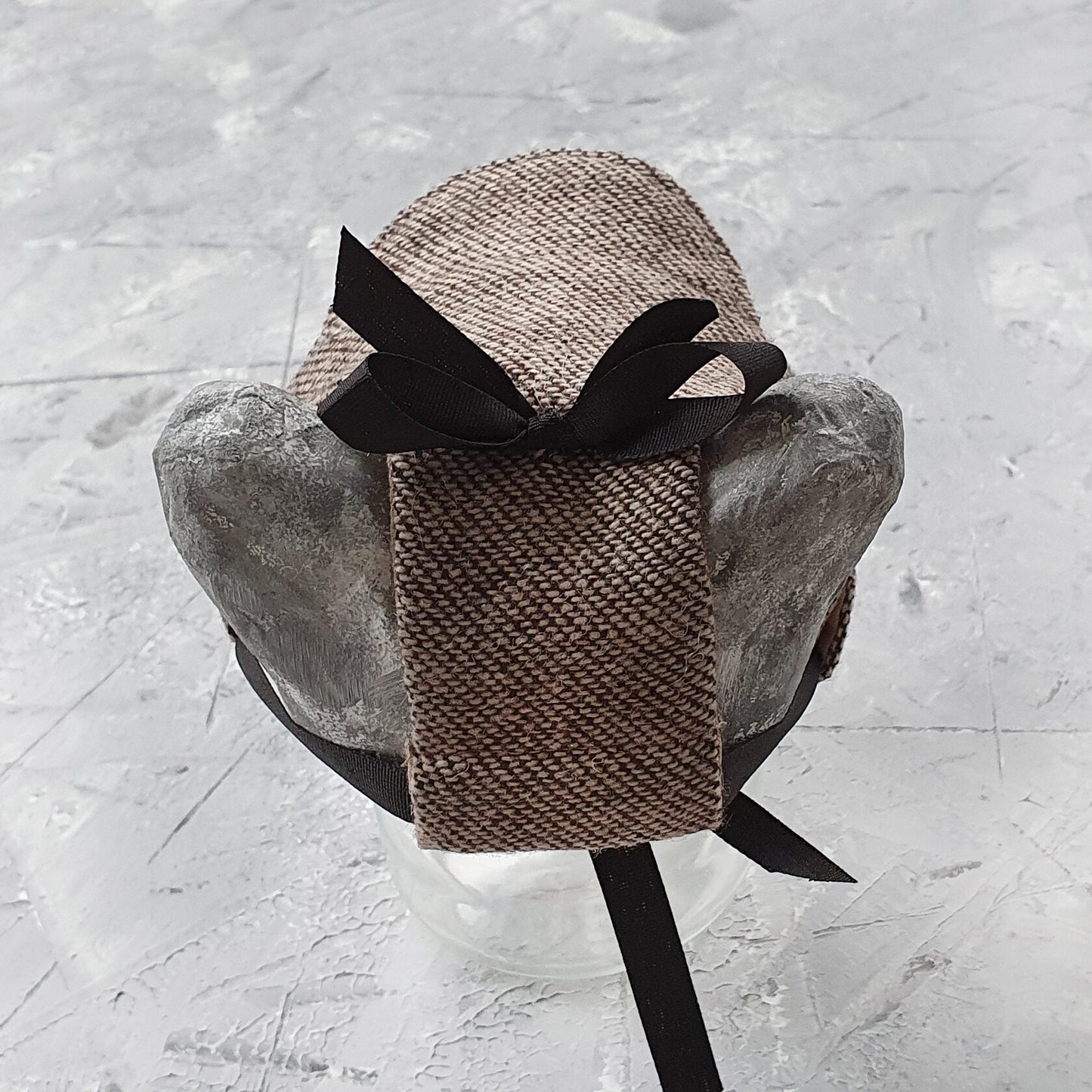 Tweed-style hat for a cat with a small brim and a black ribbon tied in a bow on top, shown on a grey textured surface.