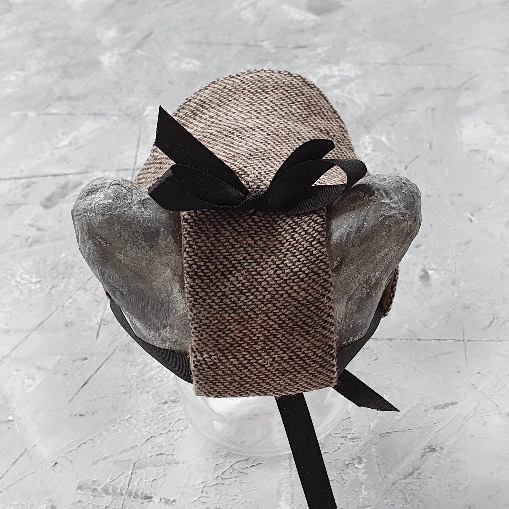 Tweed-style hat for a cat with a small brim and a black ribbon tied in a bow on top, shown on a grey textured surface.