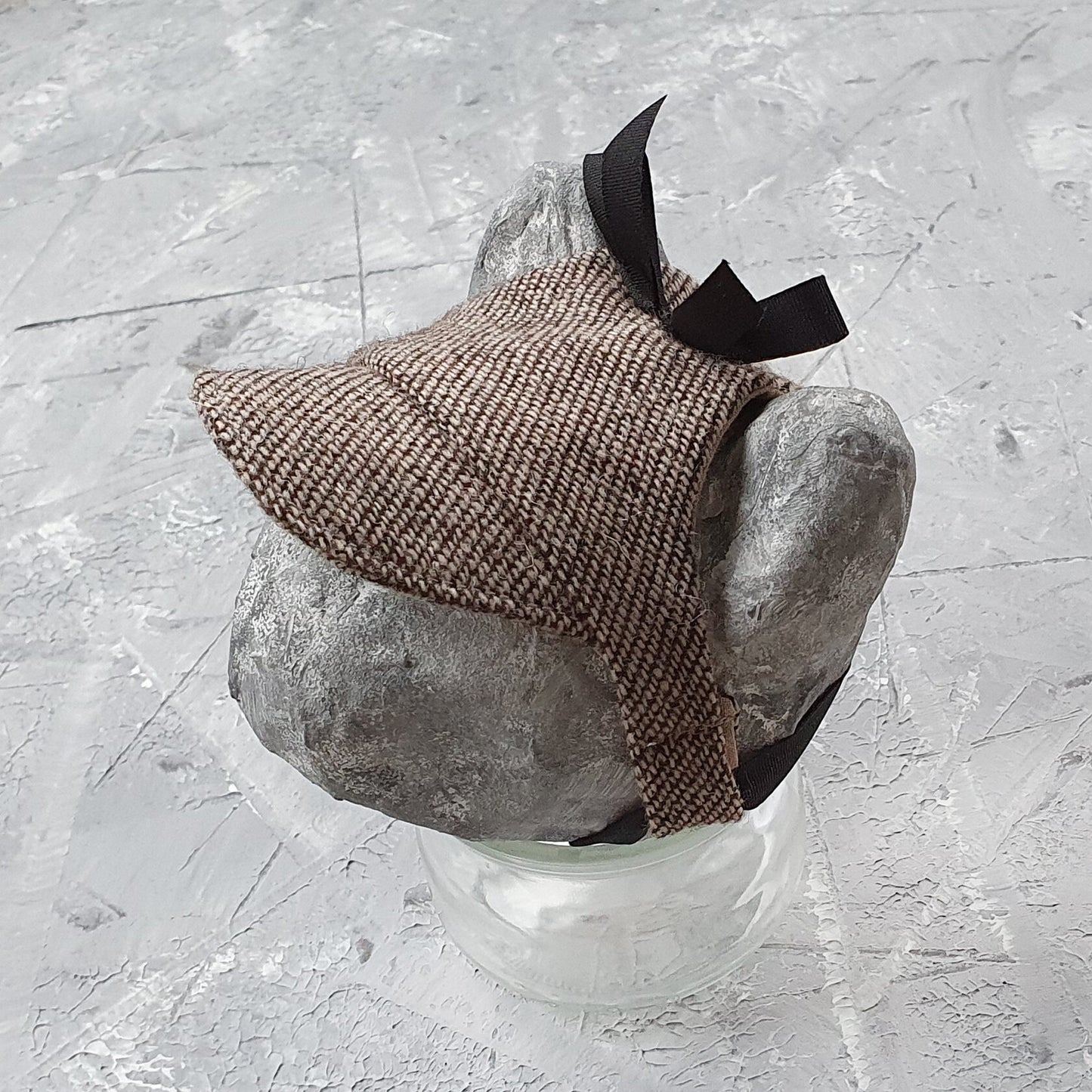 Tweed-style hat for a cat with a small brim and a black ribbon tied in a bow on top, shown on a grey textured surface.