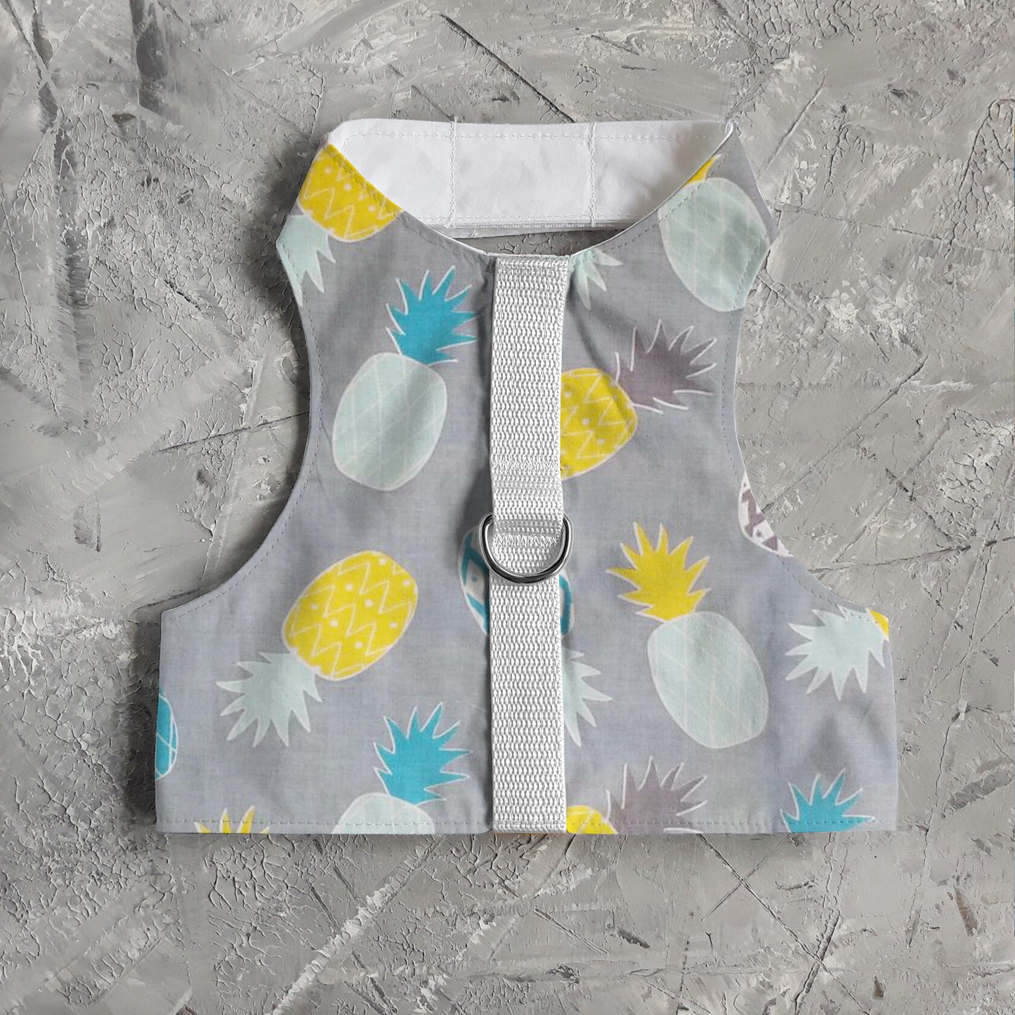 A cat harness with a repeating pineapple pattern, a D-ring for leash attachment, and adjustable straps.