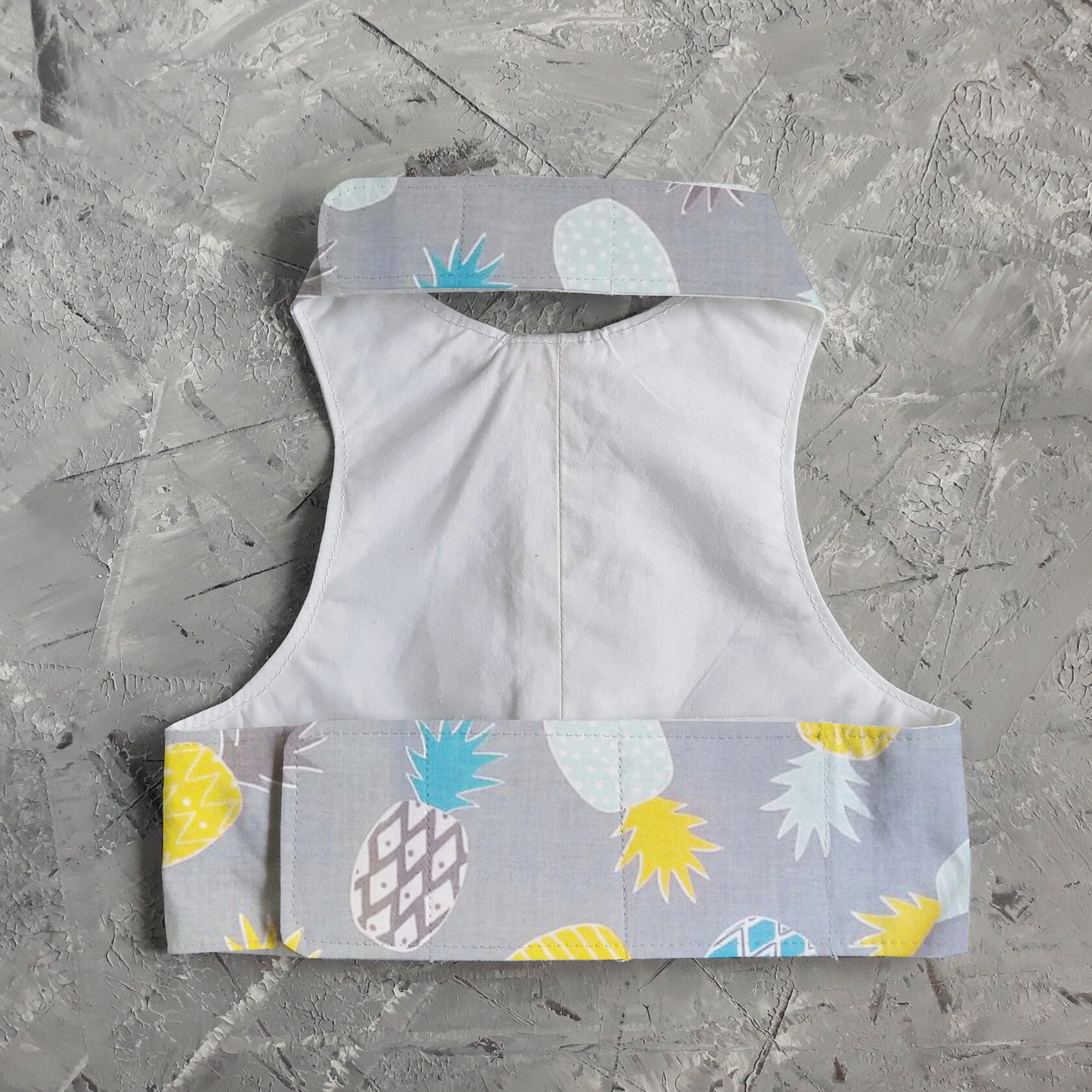 A cat harness with a repeating pineapple pattern, a D-ring for leash attachment, and adjustable straps.