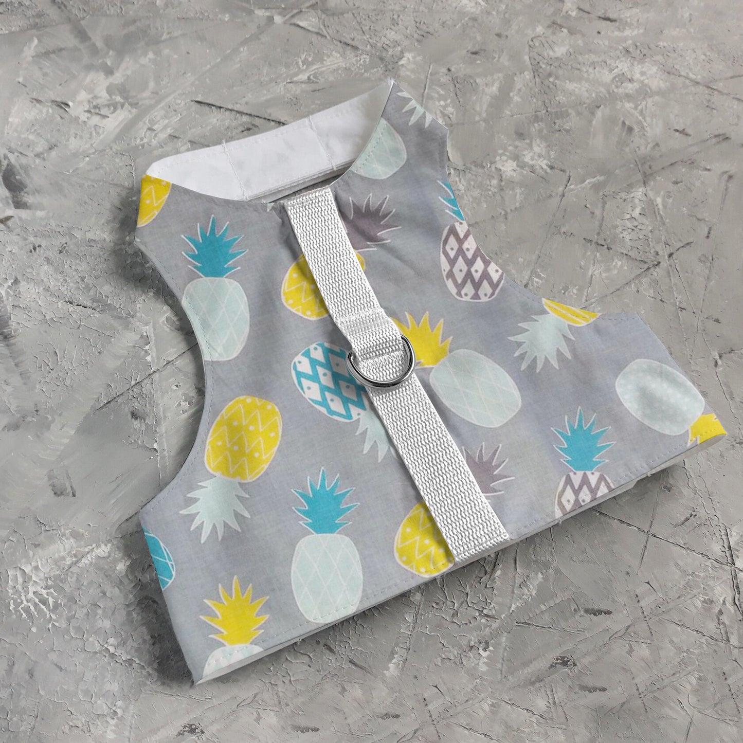 A cat harness with a repeating pineapple pattern, a D-ring for leash attachment, and adjustable straps.