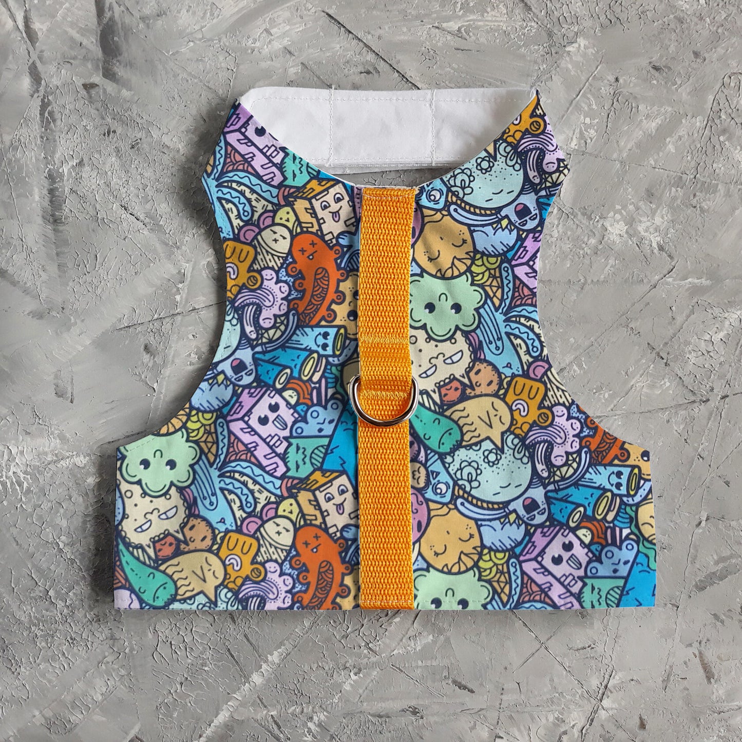 A cat harness with a colorful pattern of cartoon-like creatures and objects on a light blue background, cork side panels, a yellow vertical strap, and a silver D-ring.
