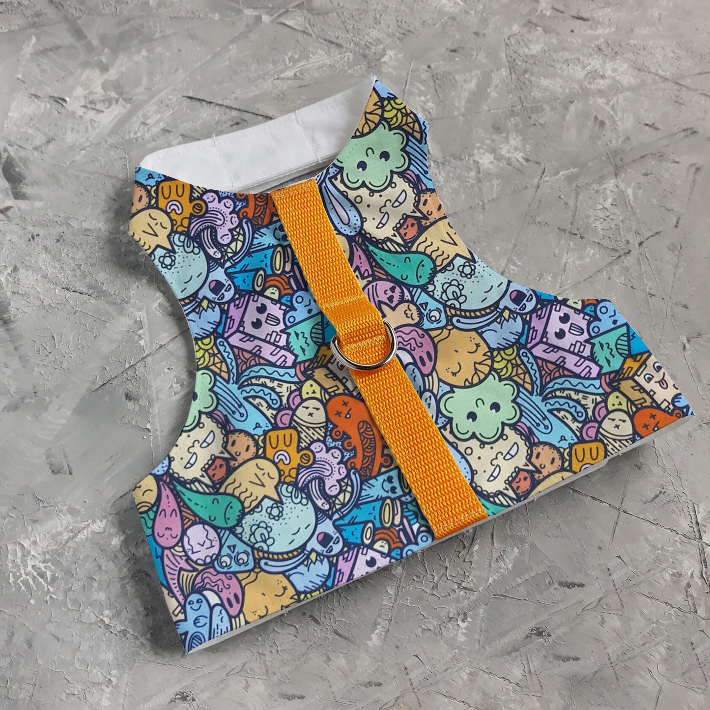 A cat harness with a colorful pattern of cartoon-like creatures and objects on a light blue background, cork side panels, a yellow vertical strap, and a silver D-ring.