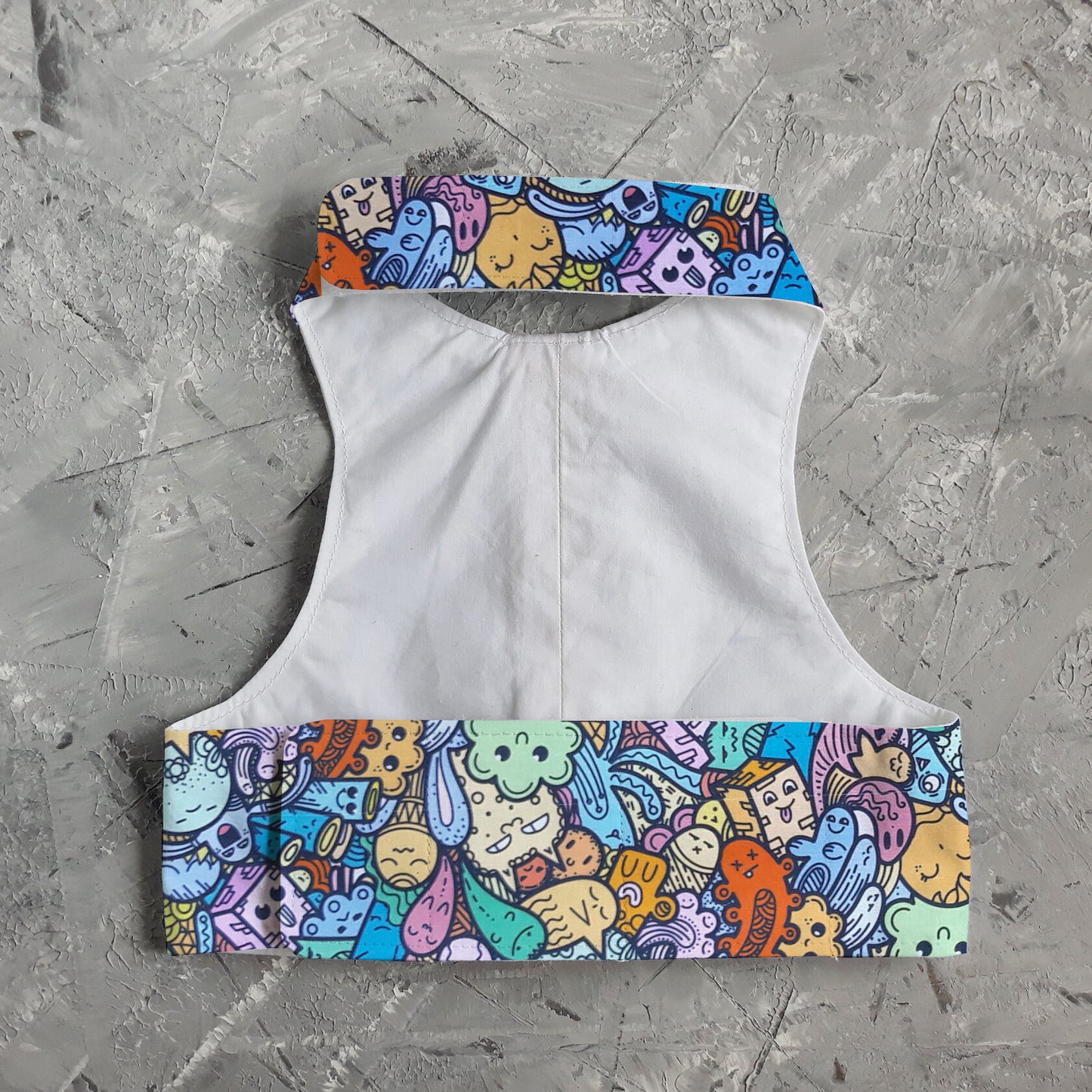 A cat harness with a colorful pattern of cartoon-like creatures and objects on a light blue background, cork side panels, a yellow vertical strap, and a silver D-ring.