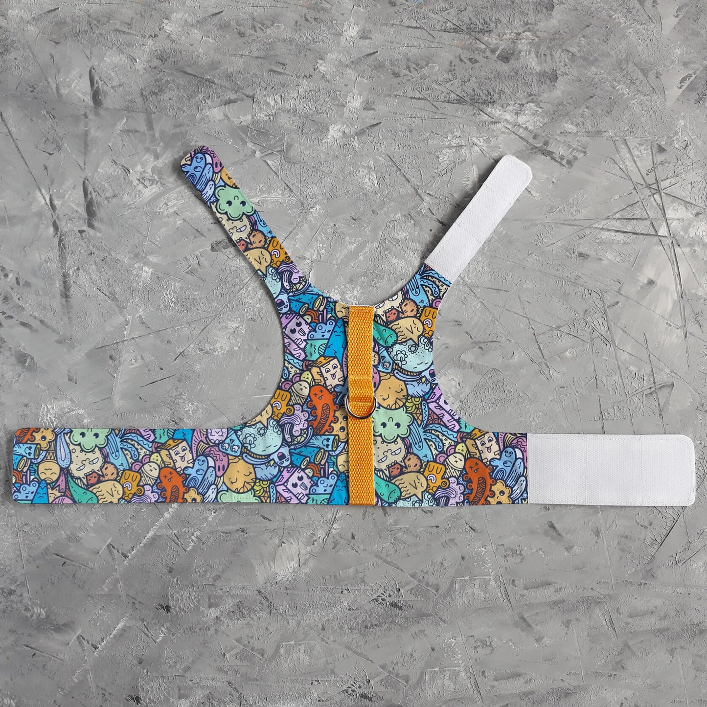A flat lay of an escape-proof and safety cat harness with a colorful pattern of cartoon-like creatures and objects, a silver D-ring, a yellow vertical strap, and white Velcro straps.