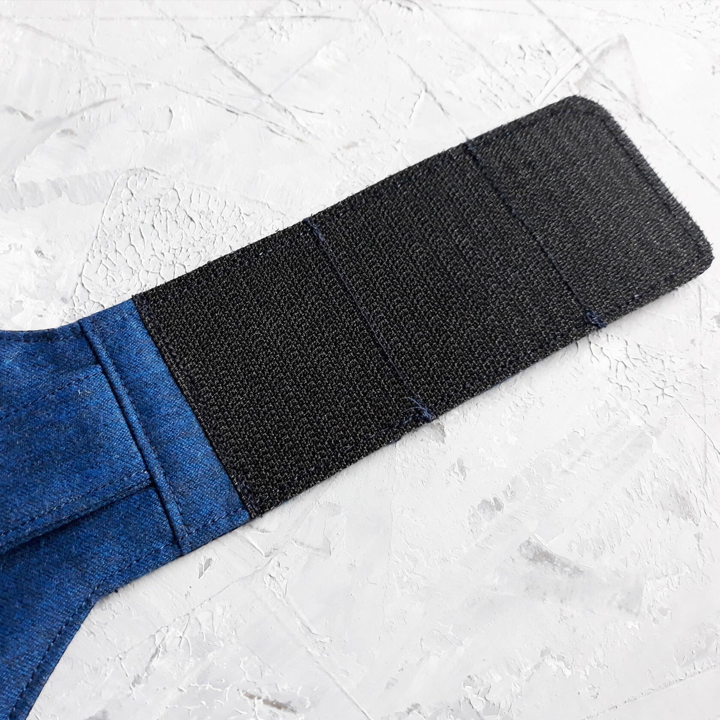 Top-down view of a blue cat harness featuring a vest design with two small pockets, silver button details, and a silver D-ring for leash attachment. Black Velcro straps extend from the sides.