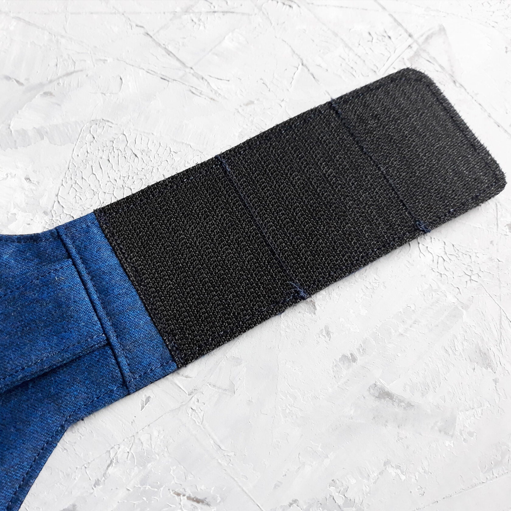 Top-down view of a blue cat harness featuring a vest design with two small pockets, silver button details, and a silver D-ring for leash attachment. Black Velcro straps extend from the sides.