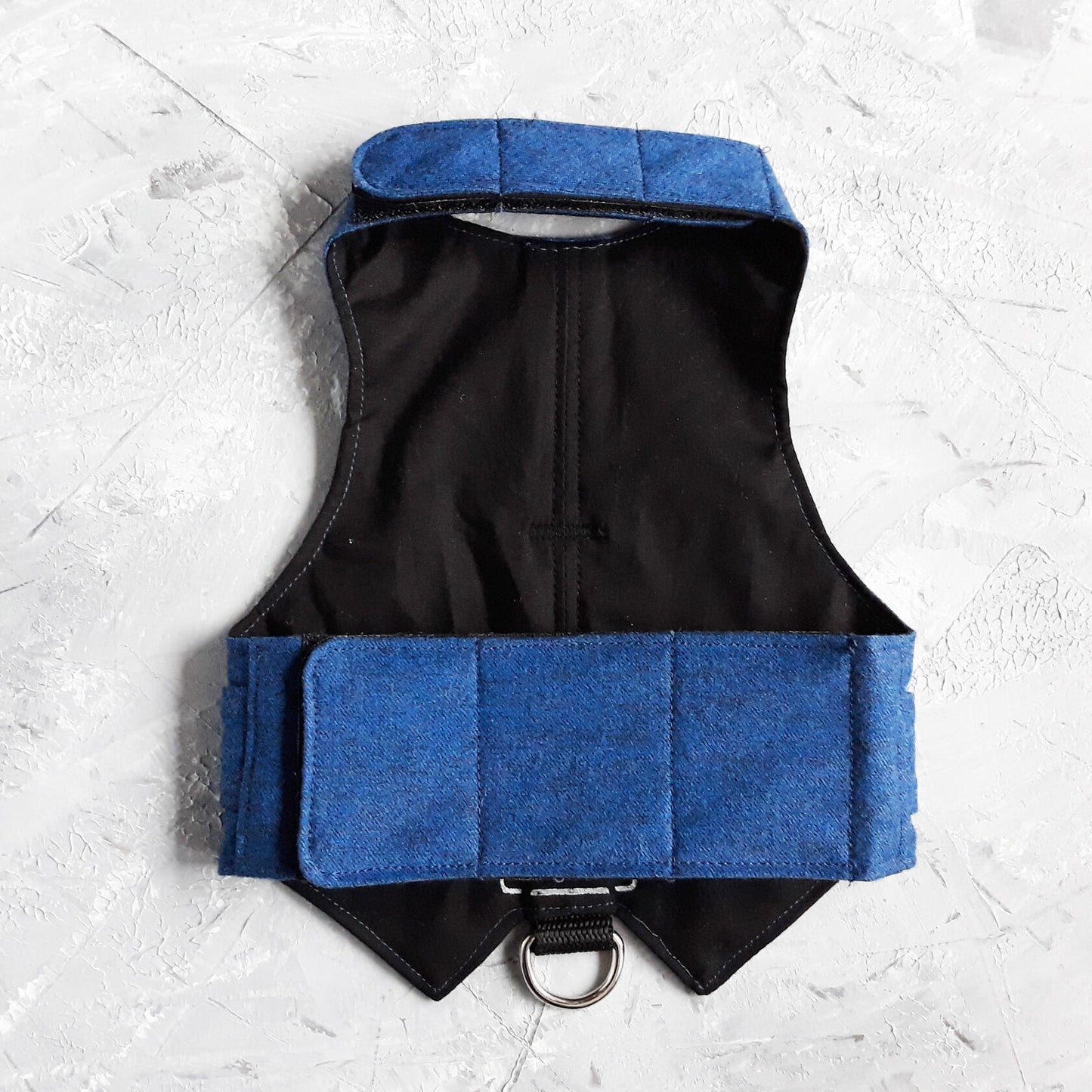 Top-down view of a blue cat harness featuring a vest design with two small pockets, silver button details, and a silver D-ring for leash attachment. Black Velcro straps extend from the sides.