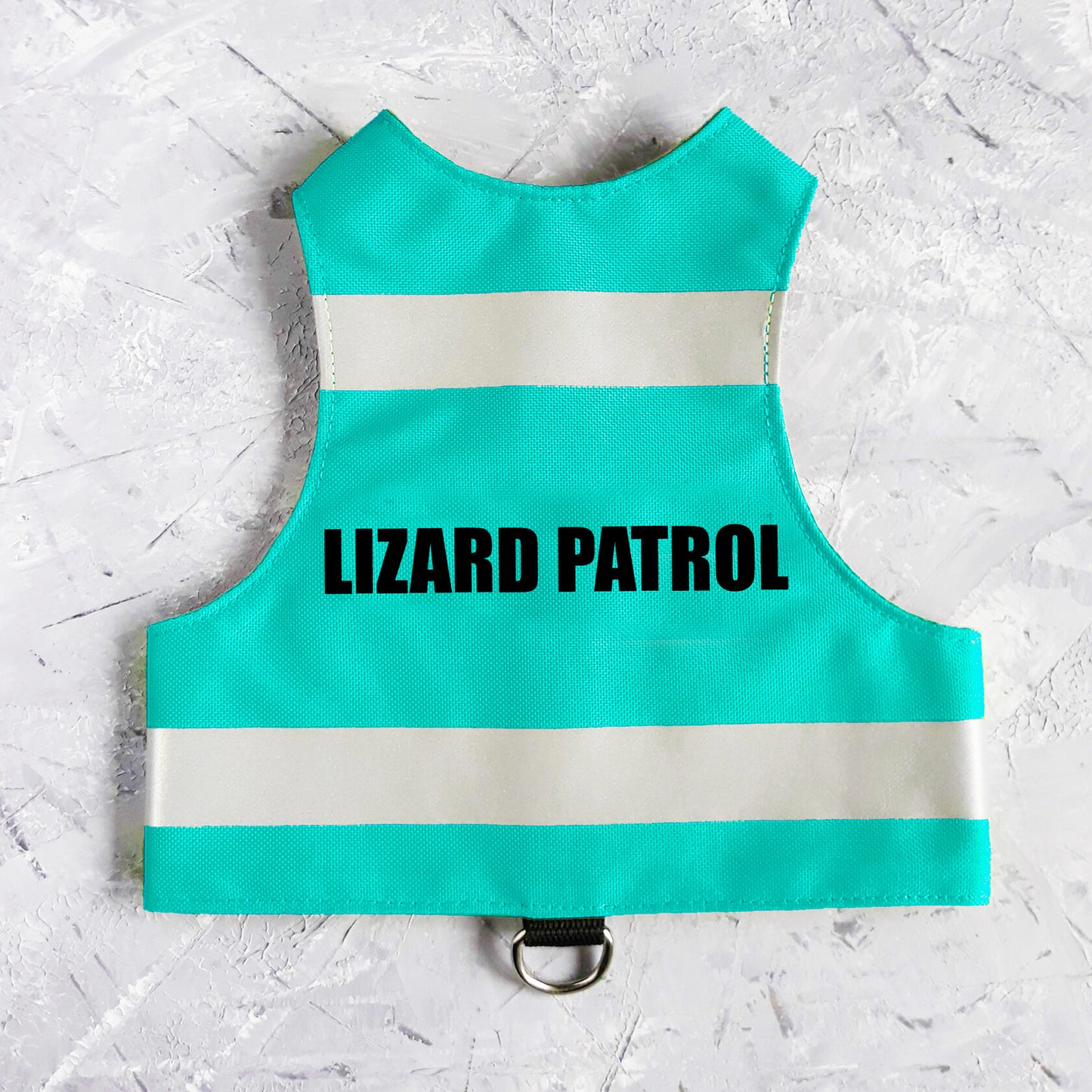Flat lay of a turquoise water-repellent safety cat harness with two horizontal reflective white stripes and the black text "LIZARD PATROL" centered on the back, featuring a black leash attachment loop with a silver D-ring at the bottom.