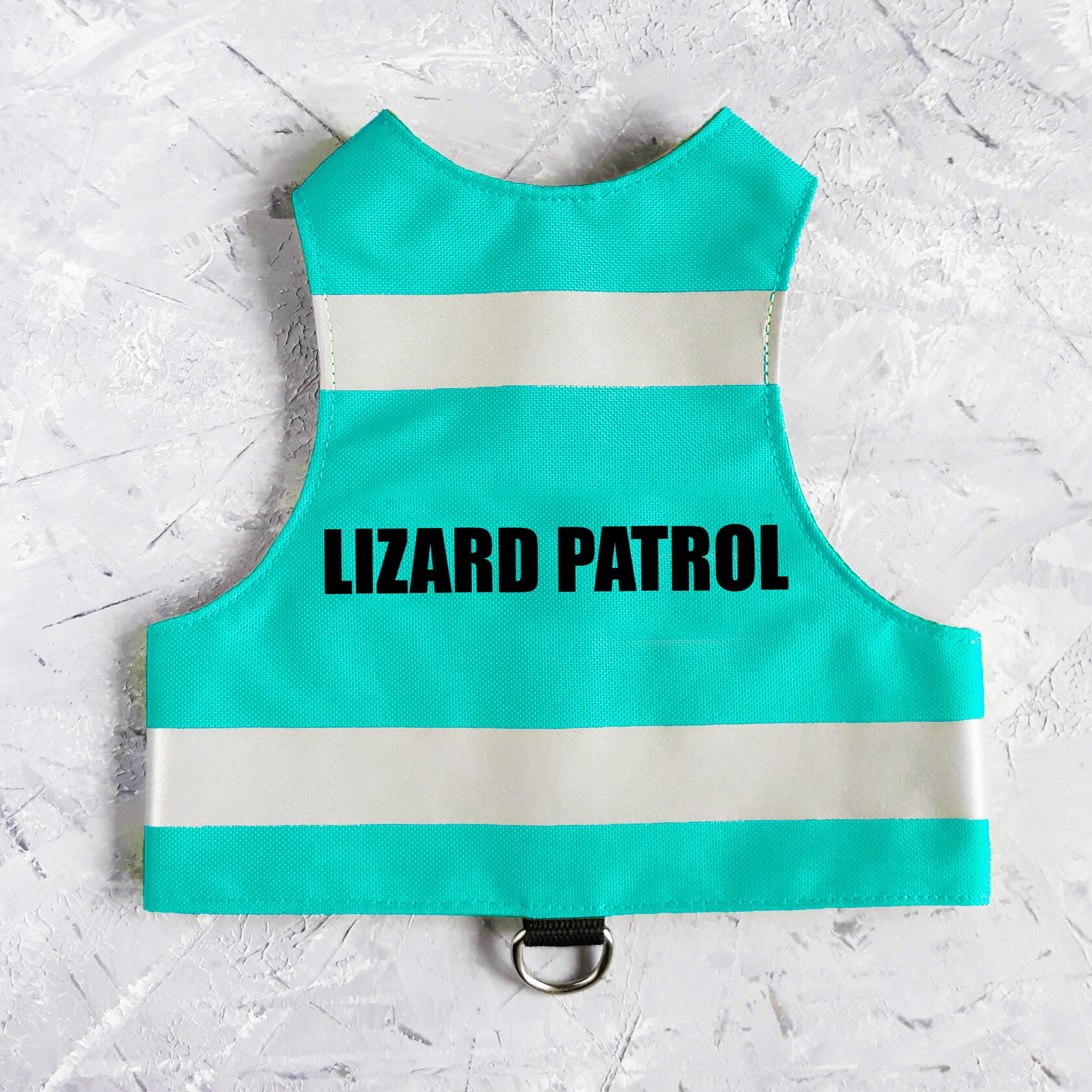 Flat lay of a turquoise water-repellent safety cat harness with two horizontal reflective white stripes and the black text "LIZARD PATROL" centered on the back, featuring a black leash attachment loop with a silver D-ring at the bottom.