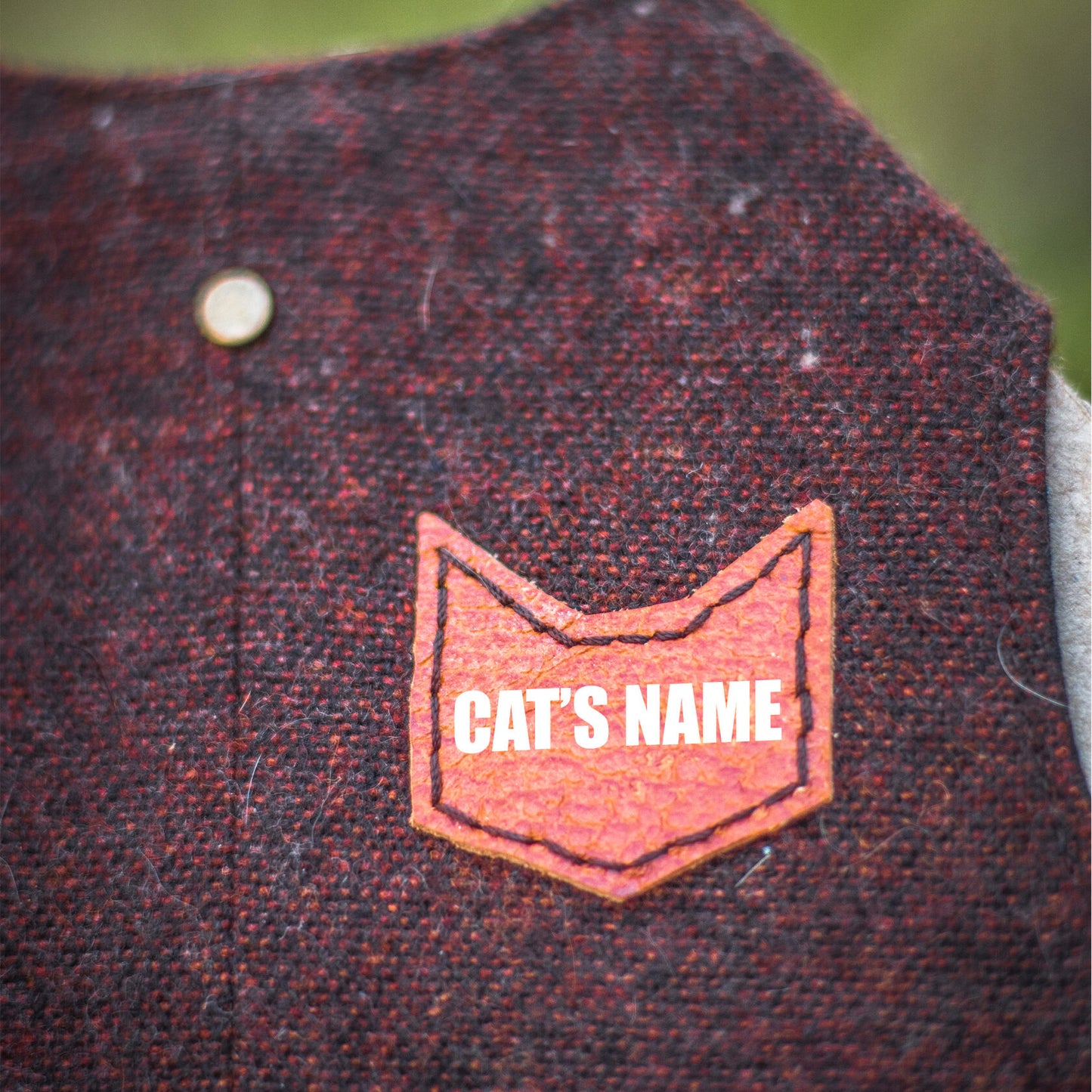 Close-up of a brown faux leather patch in the shape of a cat's head with the text "CAT'S NAME" on a burgundy tweed cat harness.