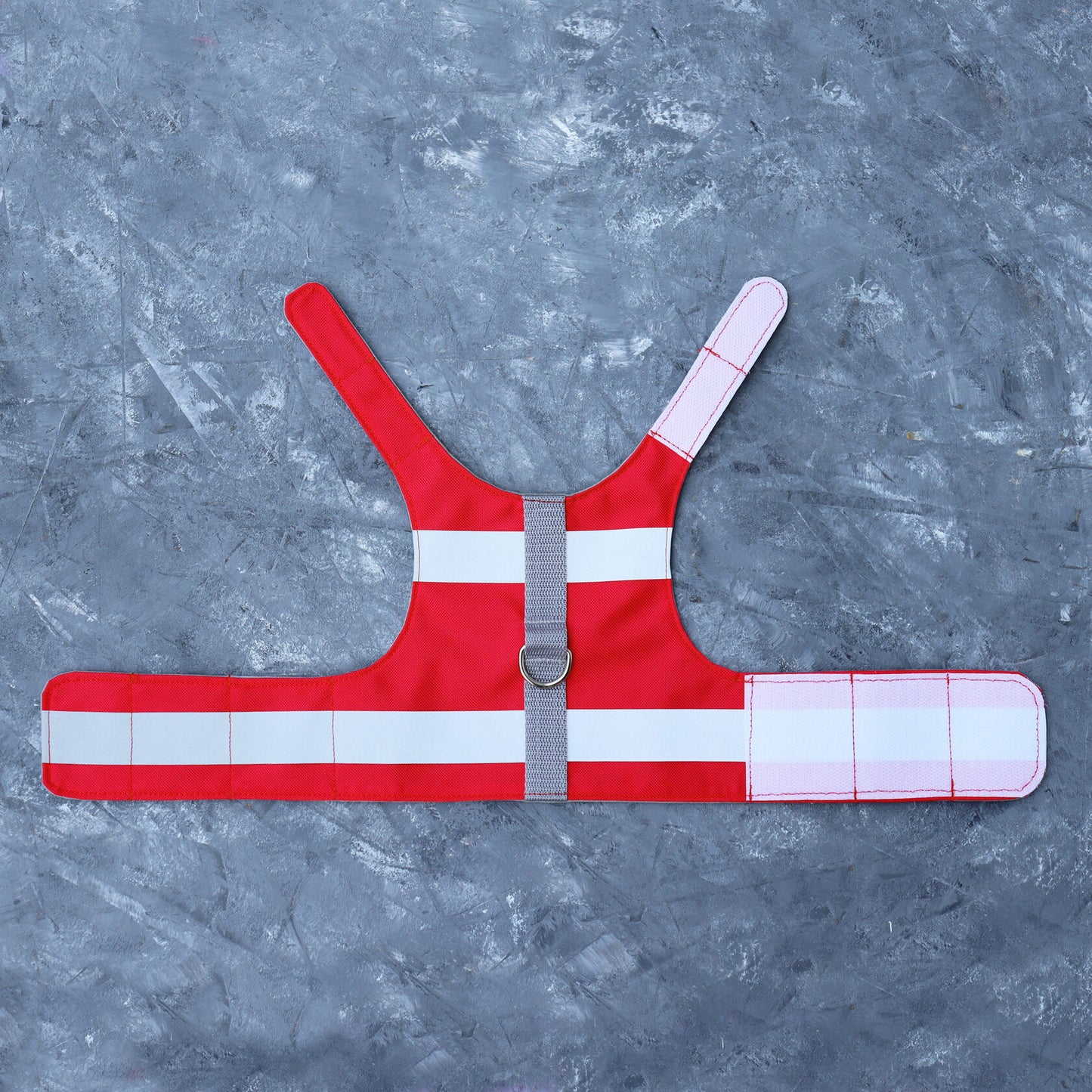 A flat lay of a bright red water-repellent cat harness with horizontal reflective silver stripes, a grey vertical strap, a silver D-ring, and white Velcro straps.