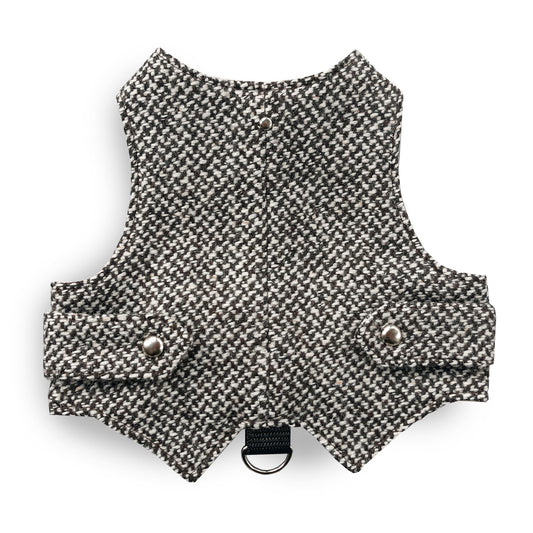 A black and white houndstooth cat harness with silver decorative buttons and a black D-ring, laid flat on a grey surface.