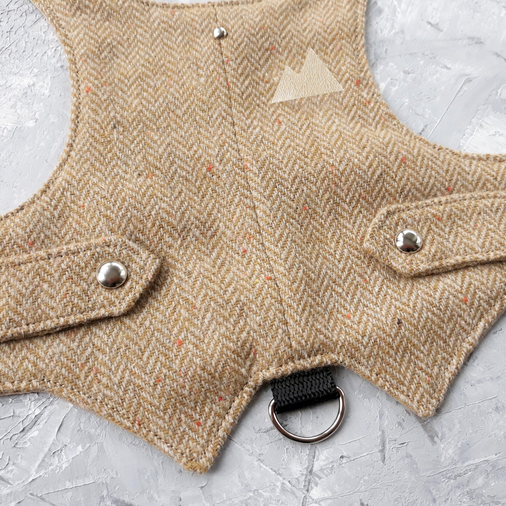 Close-up of a yellow and brown herringbone tweed cat harness with a light beige leather mountain patch, silver buttons, and a black D-ring.