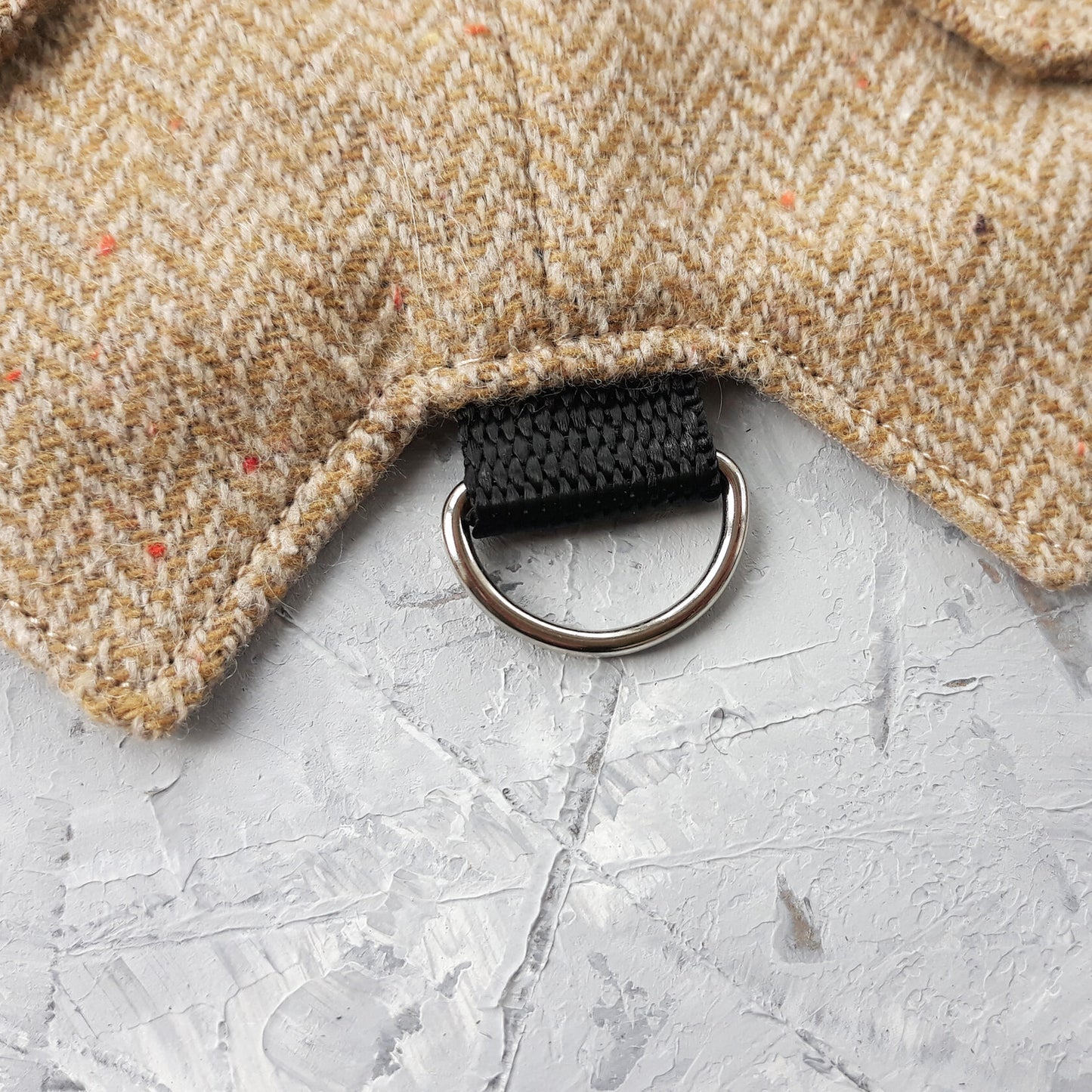 Close-up of a silver metal D-ring attached to a black strap on a yellow and brown herringbone tweed cat harness.
