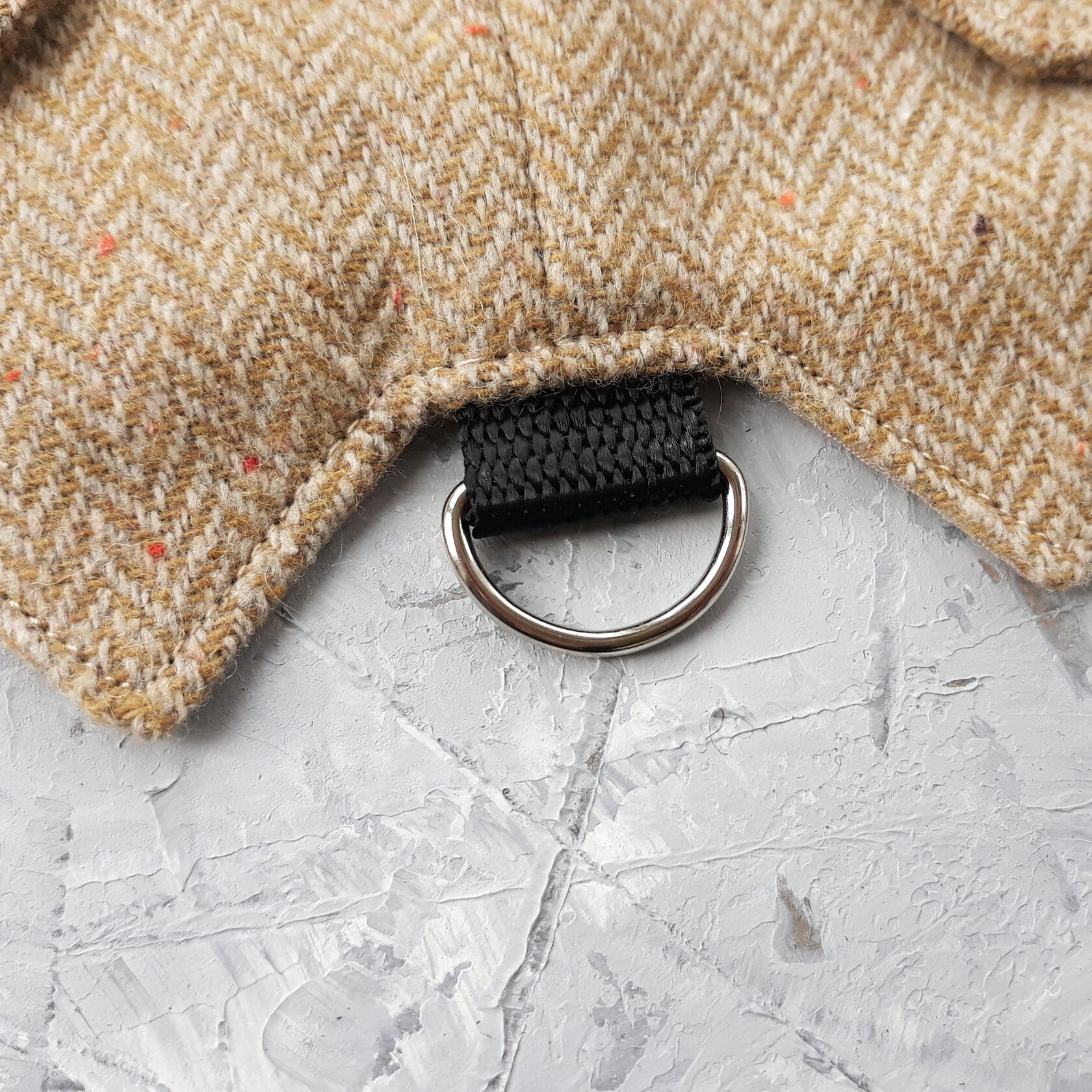 Close-up of a silver metal D-ring attached to a black strap on a yellow and brown herringbone tweed cat harness.