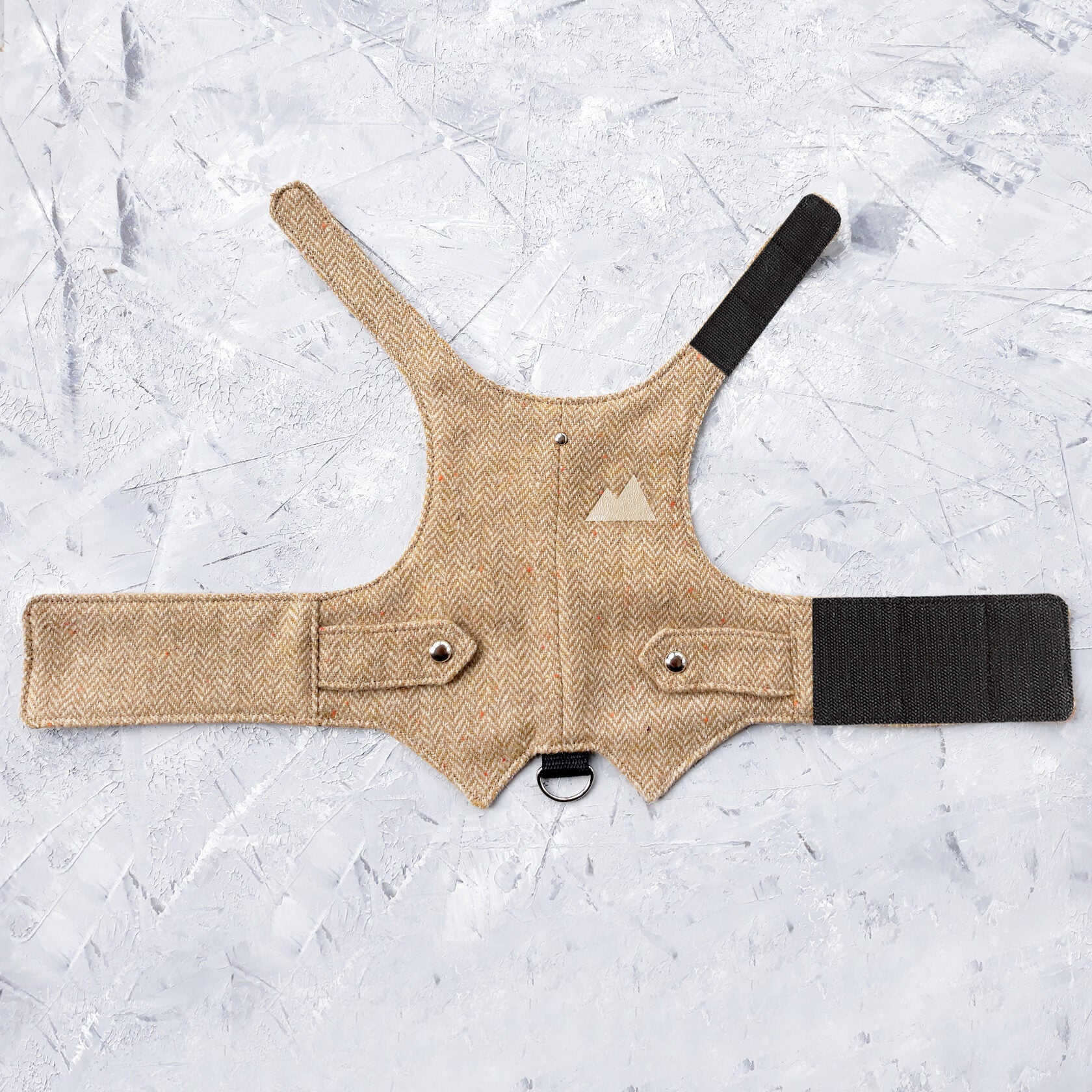 The inside of a yellow herringbone tweed cat harness showing a plain beige fabric lining, black Velcro straps, a black D-ring attachment, and a small mountain patch outline.