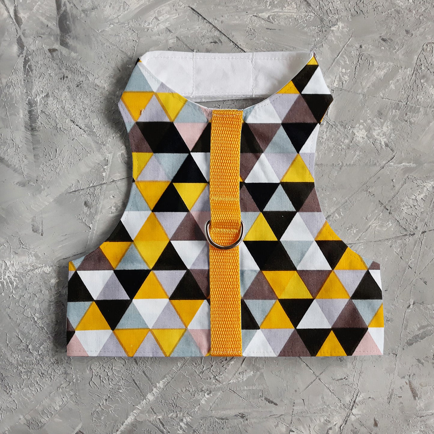 A cat harness with a geometric triangle pattern in yellow, grey, white, and black, a silver D-ring, a yellow vertical strap.