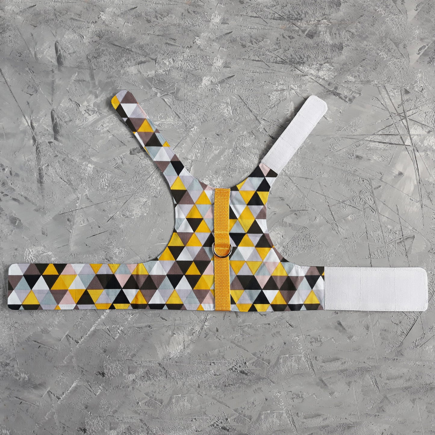 A flat lay of a cat harness with a geometric triangle pattern in yellow, grey, white, and black, a silver D-ring, a yellow vertical strap, and white Velcro straps.