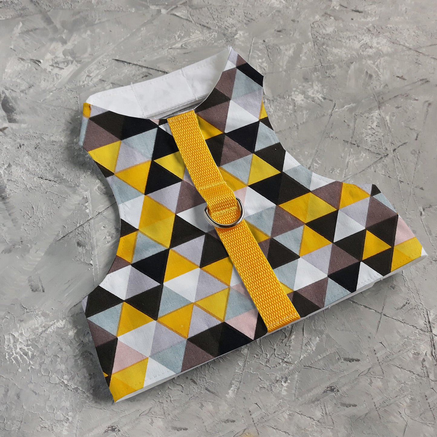 A flat lay of a cat harness with a geometric triangle pattern in yellow, grey, white, and black, a silver D-ring, and a bright yellow vertical strap.