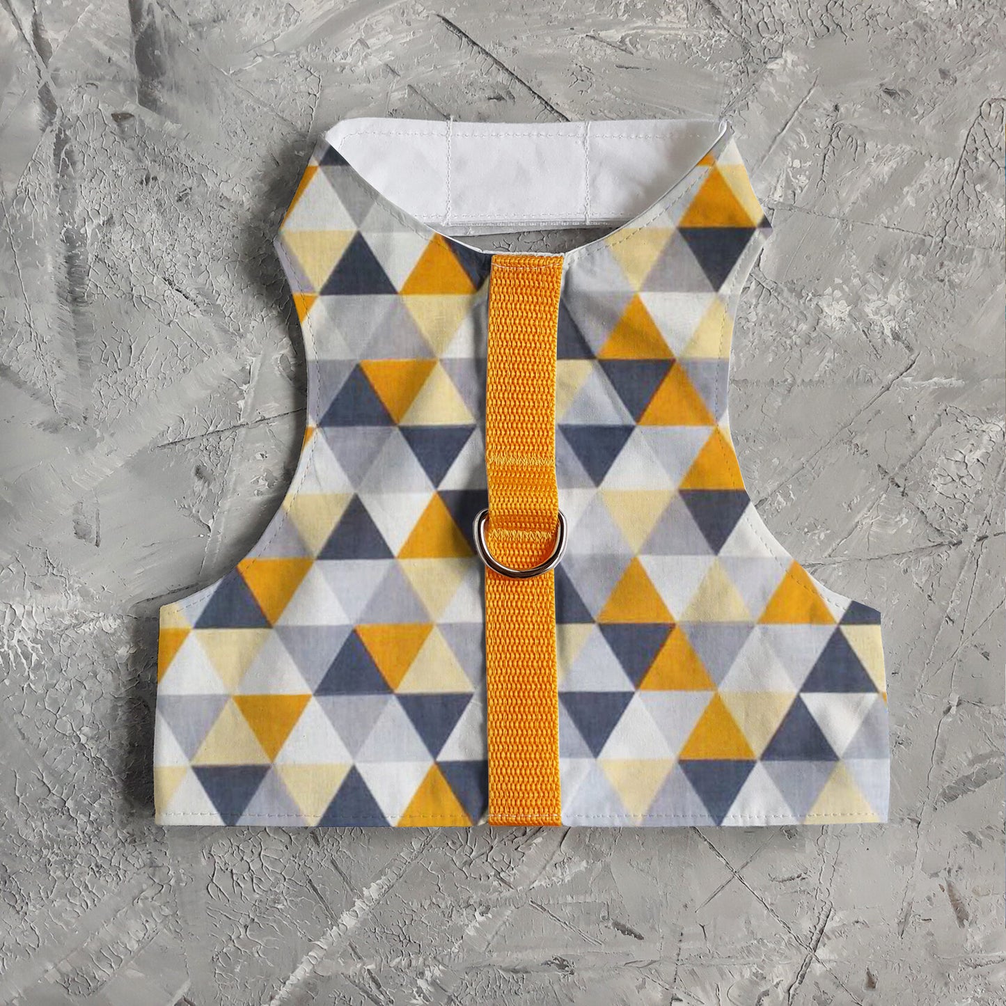 A flat lay of a safety cat harness with a geometric triangle pattern in yellow, grey, white, and black, a silver D-ring, and a bright yellow vertical strap.