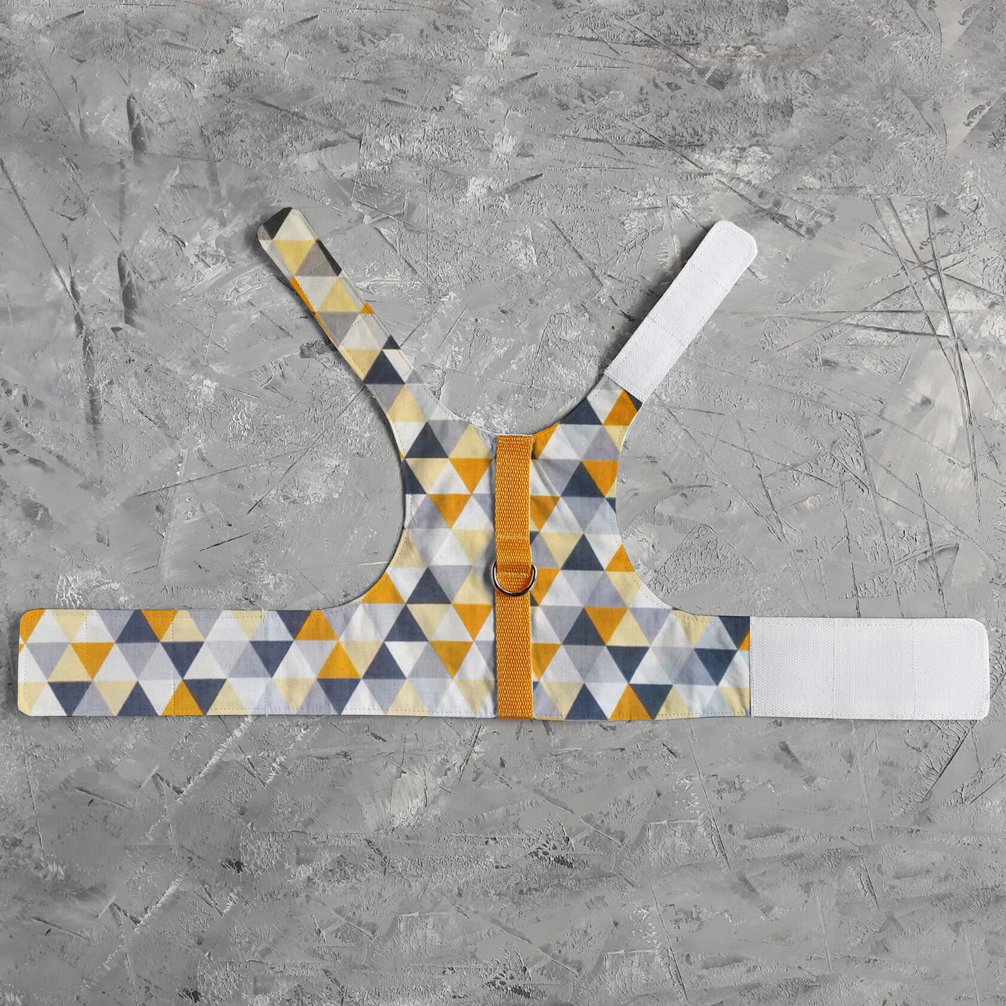 A flat lay of an escape-proof and safety cat harness with a geometric triangle pattern in yellow, grey, white, and black, a silver D-ring, a yellow vertical strap, and white Velcro straps.