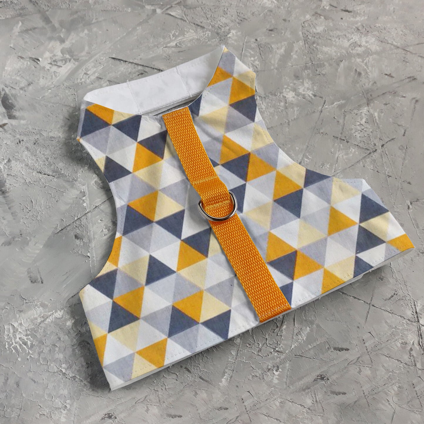 A flat lay of a safety cat harness with a geometric triangle pattern in yellow, grey, white, and black, a silver D-ring, and a bright yellow vertical strap.