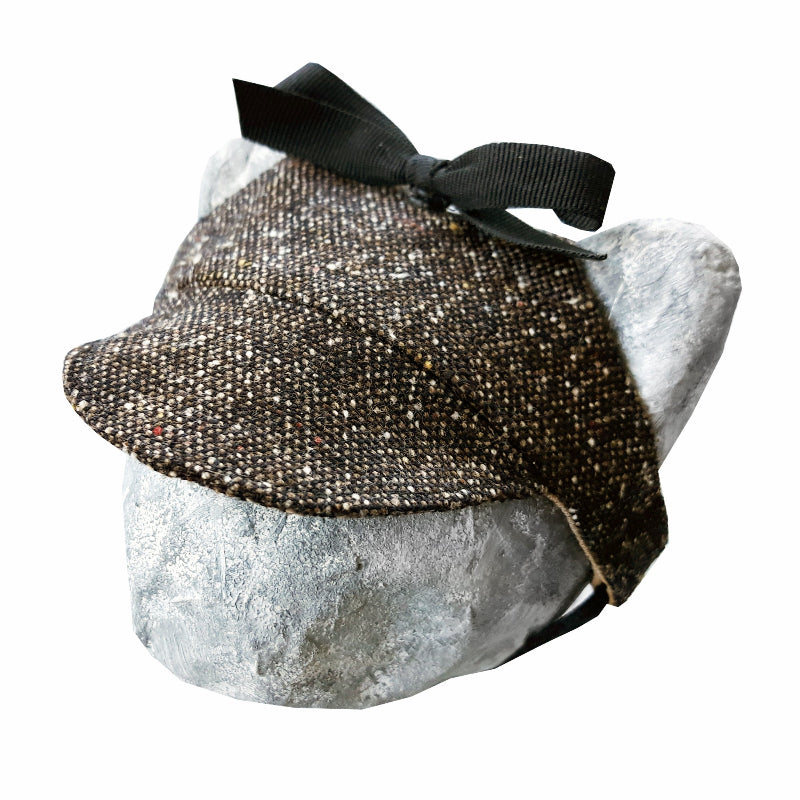 Tweed-style hat for a cat with a small brim and a black ribbon tied in a bow on top, shown on a grey textured surface.