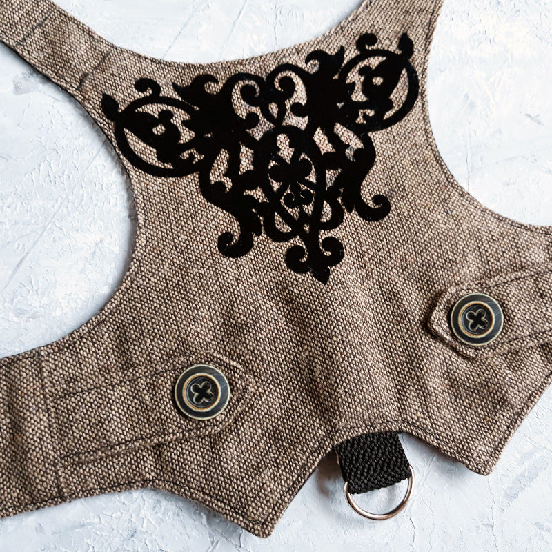 An overhead view of the inner lining of a brown tweed cat harness with a diagonal stripes pattern. The lining is a plain black fabric. A black leash attachment point with a silver D-ring is visible at the bottom.